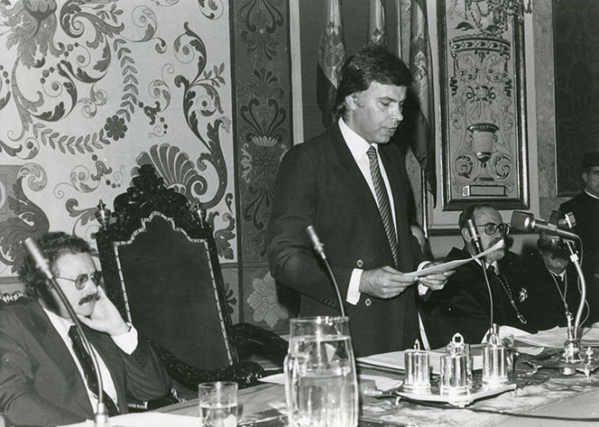 Felipe   González Márquez, Presidente del Gobierno, en pie, leyendo un discurso en el   acto de apertura del curso académico 1983/1984 de las universidades   madrileñas. A su derecha y sentado, Joaquín Leguina, Presidente de la   Comunidad de Madrid