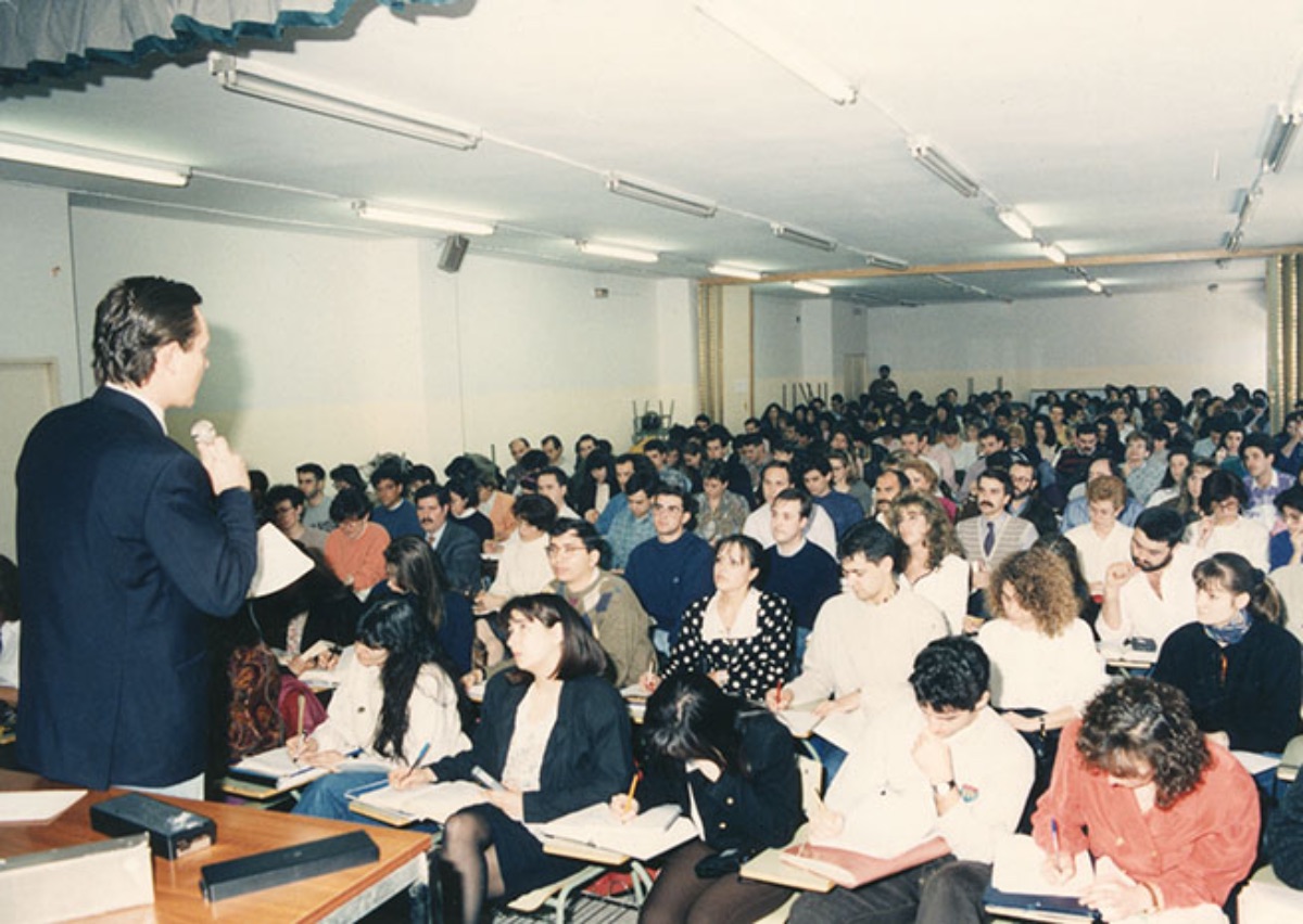 Aula   del Centro Asociado de Madrid (Barceló) llena de alumnos escuchando y tomando   apuntes durante una tutoría. El profesor, en pie, se dirige a los alumnos con   un micrófono (Imagen de Estudio fotográfico Portillo, 1993)